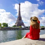 Young traveler woman in red dress and hat sitting on the quay of Seine River looking at Eiffel Tower, famous landmark and travel destination in Paris.