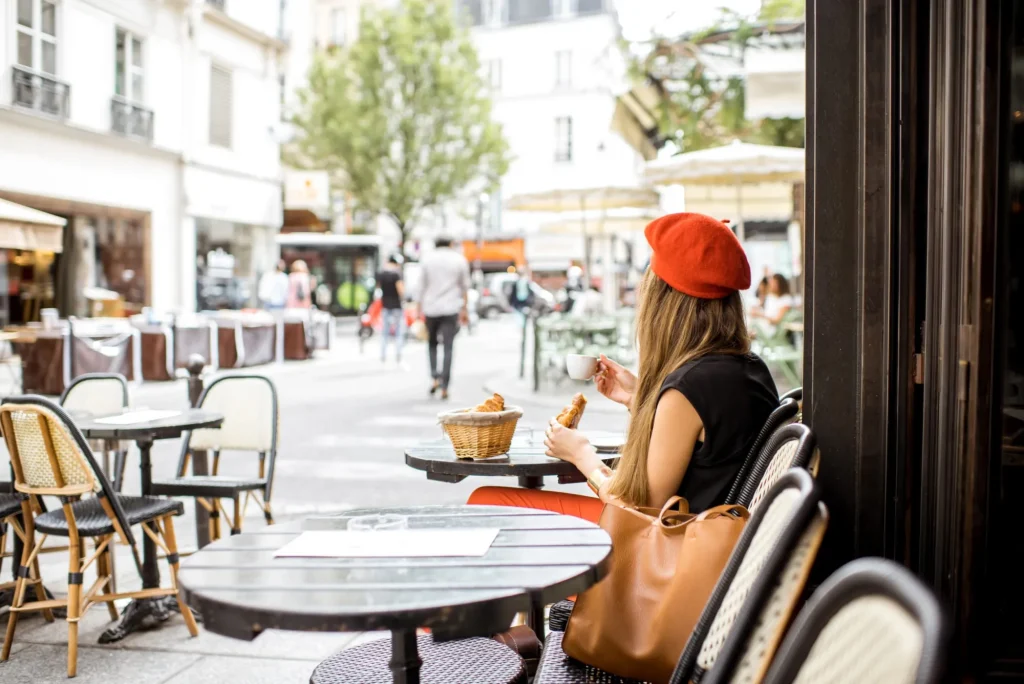 Young stylish woman in red beret having a french breakfast with coffee and croissant sitting oudoors at the cafe terrace