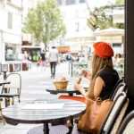 Young stylish woman in red beret having a french breakfast with coffee and croissant sitting oudoors at the cafe terrace