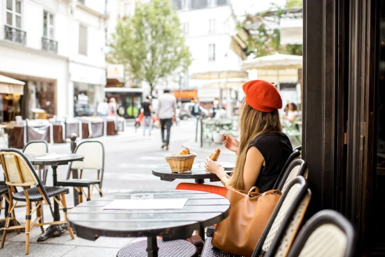 Young stylish woman in red beret having a french breakfast with coffee and croissant sitting oudoors at the cafe terrace
